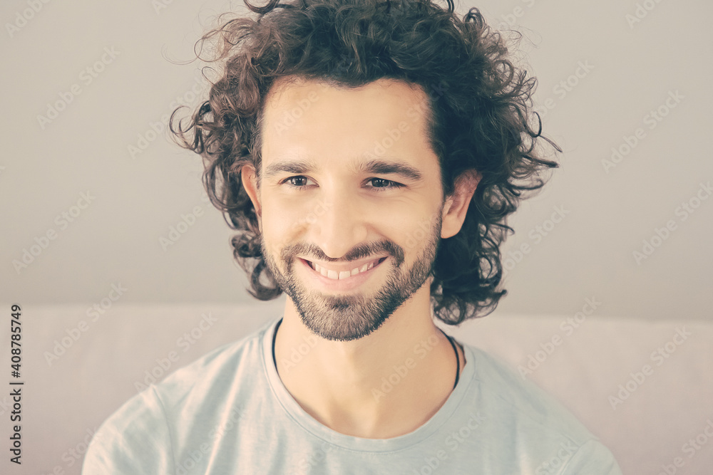 Joyful handsome curly haired guy wearing casual t-shirt, sitting on couch at home, looking away, smiling and laughing. Male portrait concept Joyful handsome curly haired guy wearing casual t-shirt, sitting on couch at home, looking away, smiling and laughing. Male portrait concept