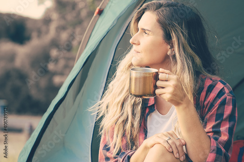 Beautiful woman holding cup of tea, sitting in tent and looking away. Caucasian female hiker relaxing on nature, enjoying and camping. Backpacking tourism, adventure and summer vacation concept