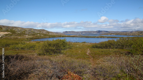 Blue lakes in the mountains. A path leading to the lakes. Green bushes and grass grow around the lakes. Stone hills are visible in the background. Beautiful white clouds float across the blue sky.