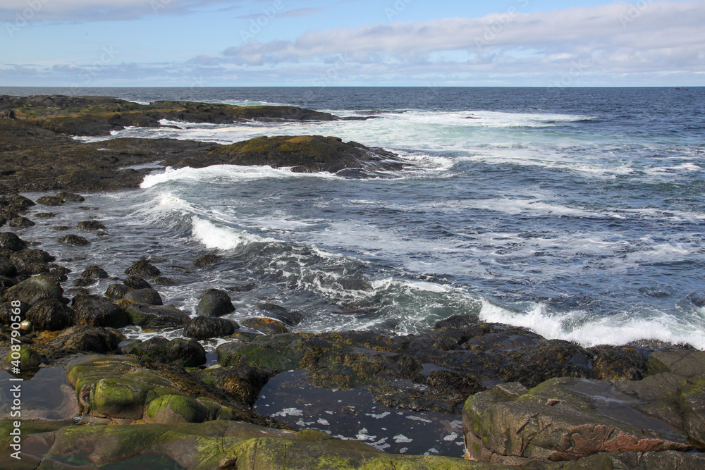 The beautiful waves of the Barents Sea break on the rocks lying on the shore. Dark blue water of the bay of the Barents Sea. Summer sea landscape. The edge of the earth. Cold water of the north sea. The beautiful waves of the Barents Sea break on the rocks lying on the shore. Dark blue water of the bay of the Barents Sea. Summer sea landscape. The edge of the earth. Cold water of the north sea.