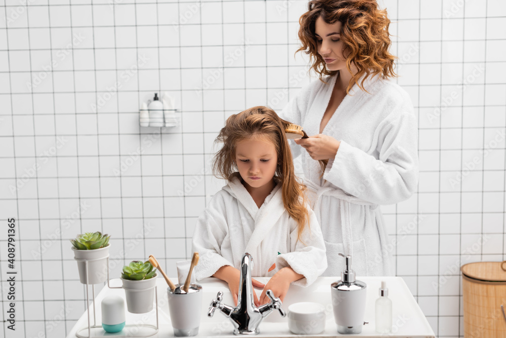 Woman brushing hair of daughter near sink in modern bathroom