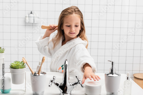 Smiling girl brushing hair and taking cream in bathroom