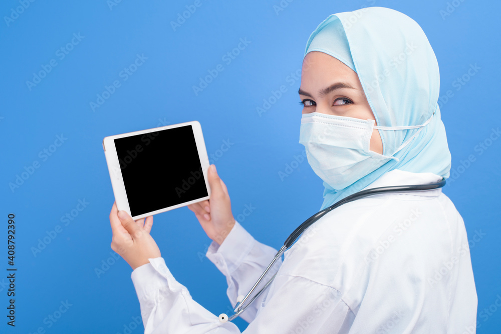 Female muslim doctor with hijab wearing a surgical mask using tablet over blue background studio. Female muslim doctor with hijab wearing a surgical mask using tablet over blue background studio.