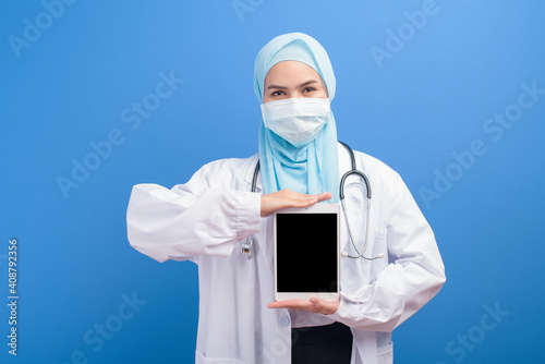 Female muslim doctor with hijab wearing a surgical mask using tablet over blue background studio.