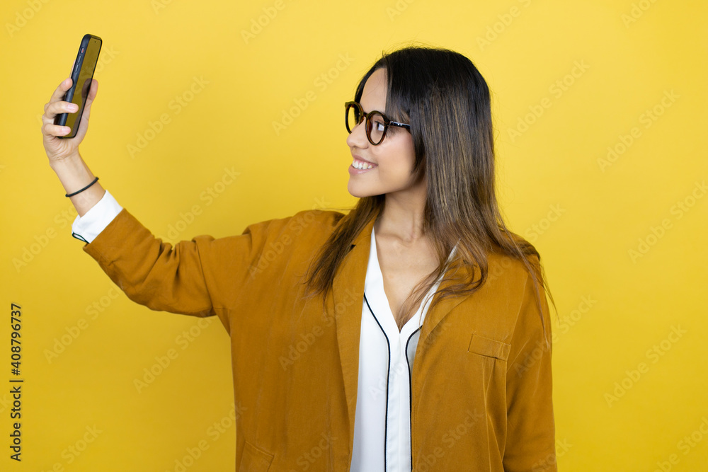 Young beautiful woman wearing a blazer over isolated yellow background taking a selfie with her phone