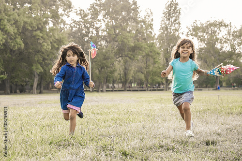 Joyful black haired little girls with pinwheels running on grass, having race in park. Front view, full length. Children outdoor activity concept