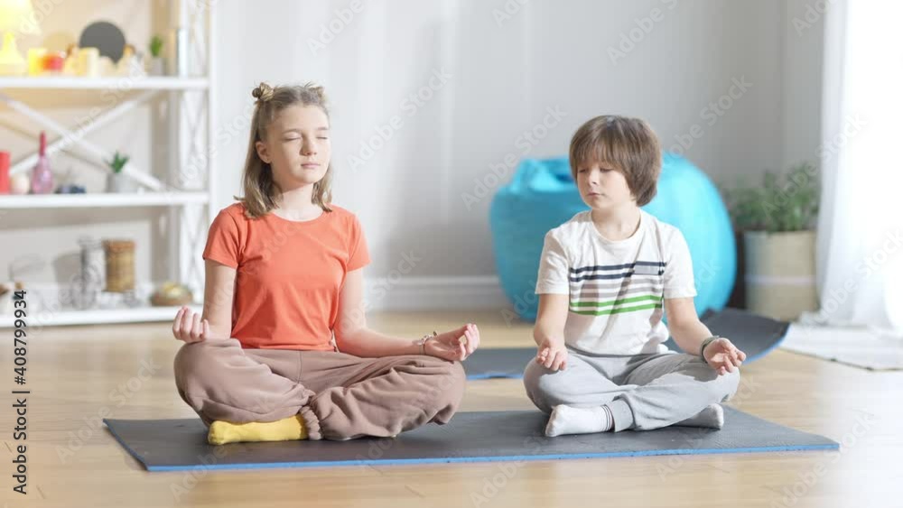 Concentrated Caucasian girl sitting on exercise mat with eyes closed as boy repeating yoga training after sibling. Wide shot of little yogi exercising indoors at home. Sport and wellbeing concept. Concentrated Caucasian girl sitting on exercise mat with eyes closed as boy repeating yoga training after sibling. Wide shot of little yogi exercising indoors at home. Sport and wellbeing concept.