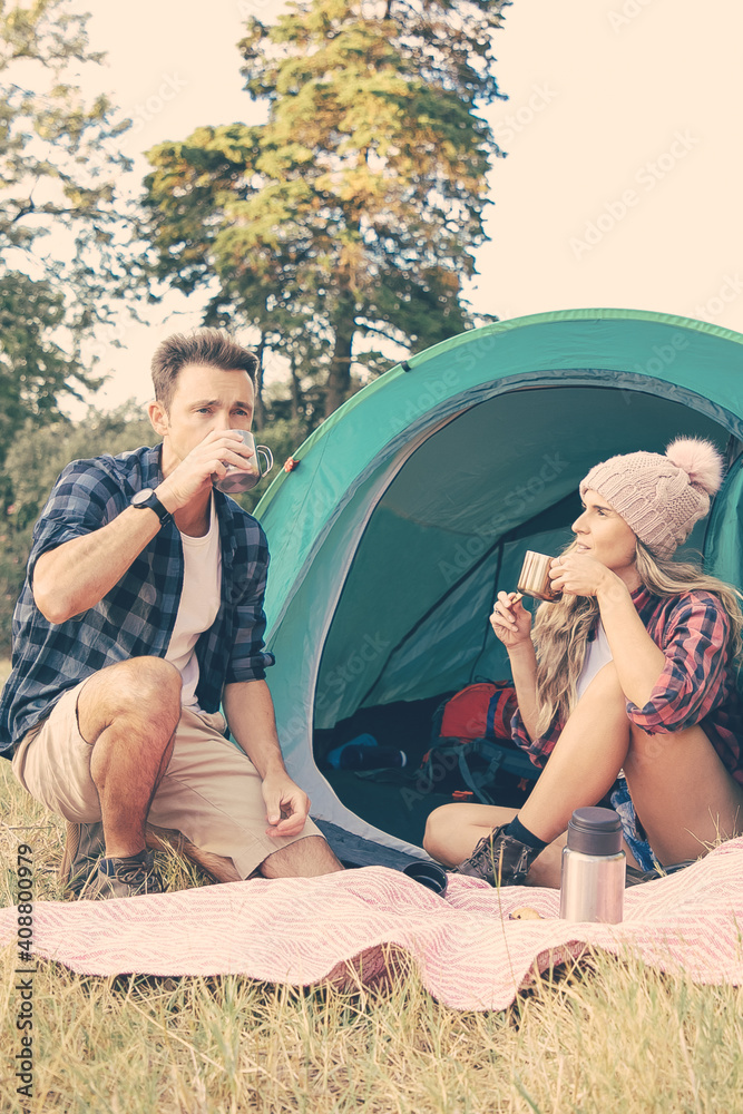 Young tourists sitting in tent and drinking tea from thermos. Caucasian travelers camping on nature and relaxing on lawn together. Backpacking tourism, adventure and summer vacation concept
