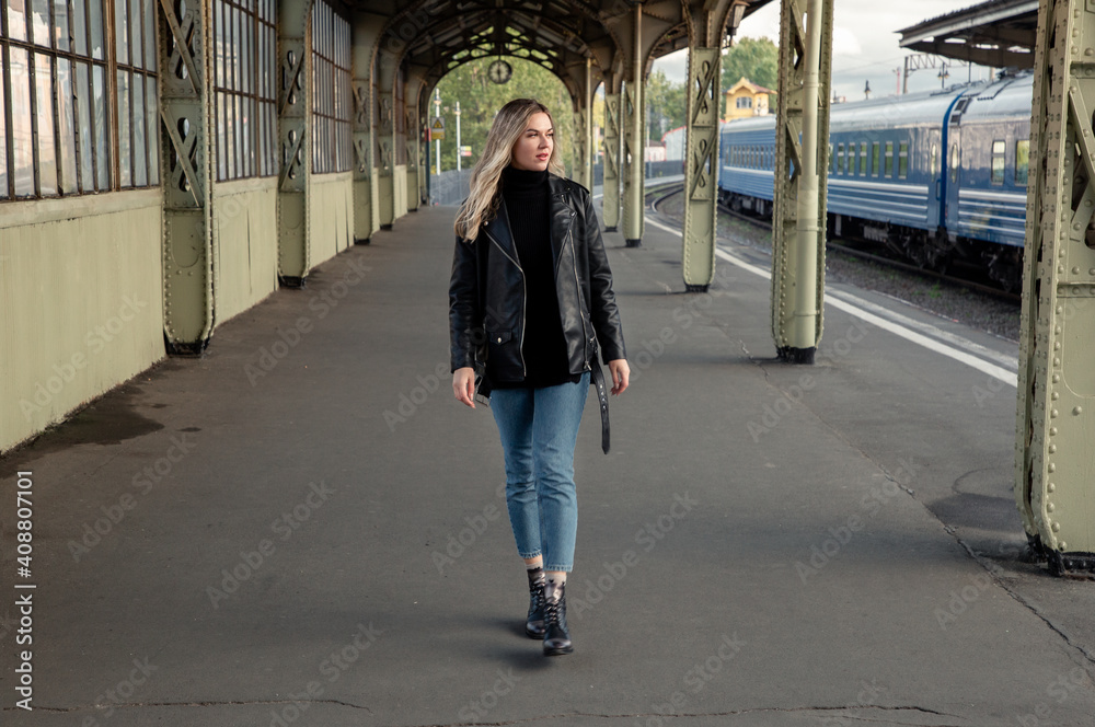A young blonde woman in a leather jacket and blue jeans walks along the platform of the old railway station