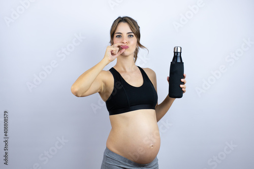 Young sportswoman pregnant wearing sportswear holding bottle with water over white background mouth and lips shut as zip with fingers. Secret and silent, taboo talking photo