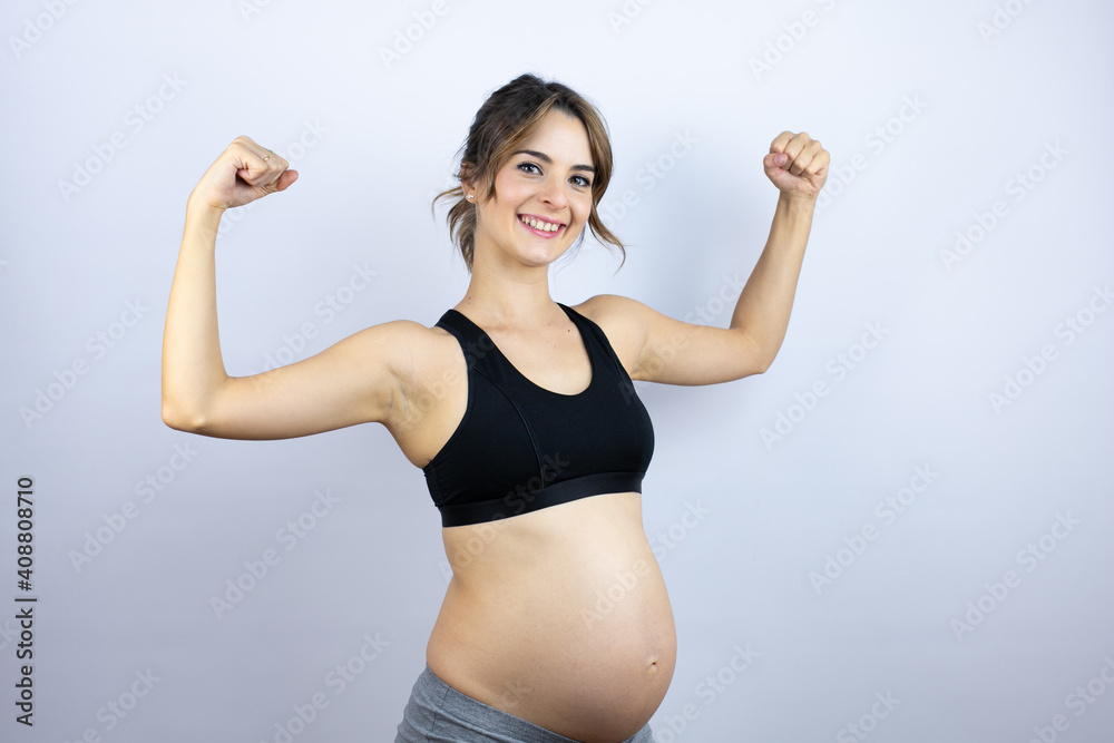 Young sportswoman pregnant wearing sportswear over white background feeling happy, satisfied and powerful, flexing fit and muscular biceps, looking strong after the gym Young sportswoman pregnant wearing sportswear over white background feeling happy, satisfied and powerful, flexing fit and muscular biceps, looking strong after the gym