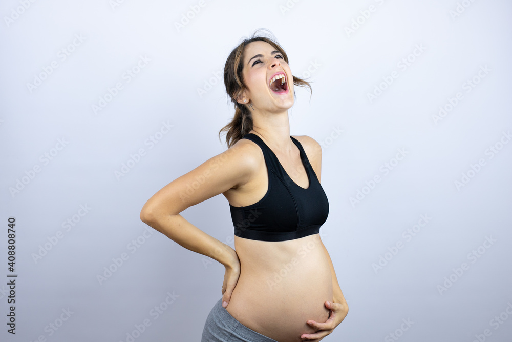 Young sportswoman pregnant wearing sportswear over white background suffering of backache, touching back with hand, muscular pain Young sportswoman pregnant wearing sportswear over white background suffering of backache, touching back with hand, muscular pain