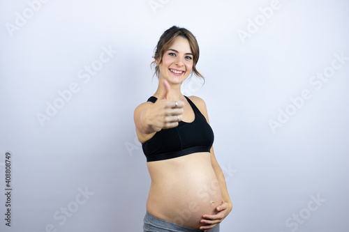 Young sportswoman pregnant wearing sportswear over white background success sign doing positive gesture with hand, thumb up smiling and happy. cheerful expression