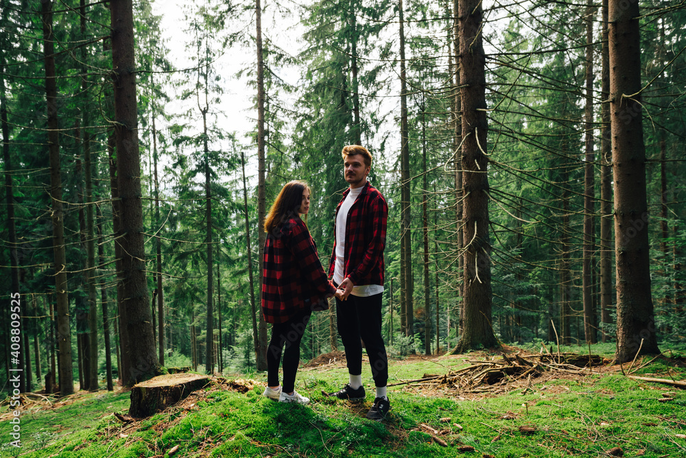 Beautiful couple man and woman in red shirts stand on the moss in the mountain forest during a walk and hold hands on a background of beautiful coniferous trees. Beautiful couple man and woman in red shirts stand on the moss in the mountain forest during a walk and hold hands on a background of beautiful coniferous trees.