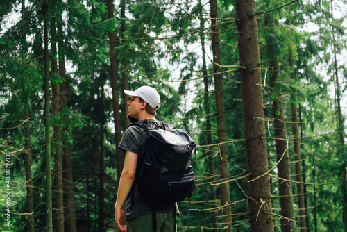 A young hipster man in casual clothes calls on the phone on a mountain hike, stands in the woods, communicates on a smartphone and looks away, carries a backpack on his shoulders and a cap