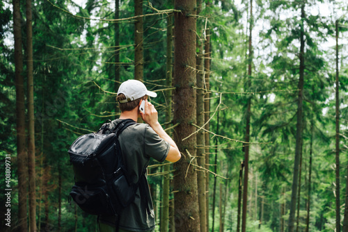 Male tourist in casual clothes and a backpack on his back communicates on a smartphone in the mountains, stands in the woods and rests on the call.