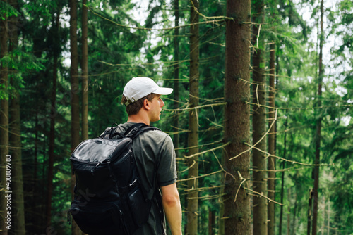 Young male hiker stands with his back in the woods with a backpack on his back and looks away with a serious face. tourist is resting on a hike.