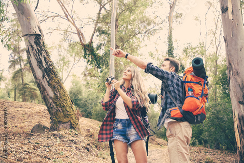 Couple hiking in forest with backpacks and camera. Attractive Caucasian man pointing at something. Blonde woman looking at it and holding camera. Tourism, adventure and summer vacation concept