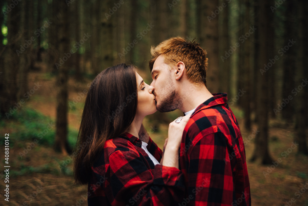 Portrait of a beautiful young man and woman kissing in the woods on a background of coniferous tree trunks. Love story photo, a couple in red shirts hugging and kissing on a hike. Portrait of a beautiful young man and woman kissing in the woods on a background of coniferous tree trunks. Love story photo, a couple in red shirts hugging and kissing on a hike.