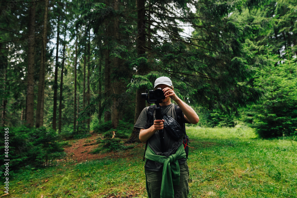 A male videographer with a camera on a stabilizer stands in the woods against a background of trees and adjusts the technique for shooting. Creating video content in nature A male videographer with a camera on a stabilizer stands in the woods against a background of trees and adjusts the technique for shooting. Creating video content in nature