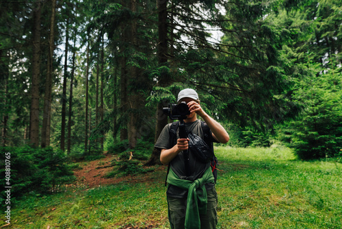 A male videographer with a camera on a stabilizer stands in the woods against a background of trees and adjusts the technique for shooting. Creating video content in nature