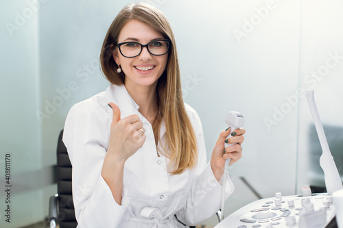 Close up portrait of professional smiling young woman doctor sonographer sitting near modern ultrasound scanner machine and holding transducer, showing her thumb up photo