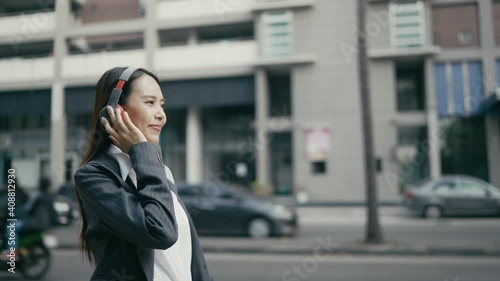  Asian woman, businesswoman, beautiful eyes, white skin, black suit, red wireless headphones. On mobile, smartphone Walking listening to music with pleasure On the side of the street in the city
