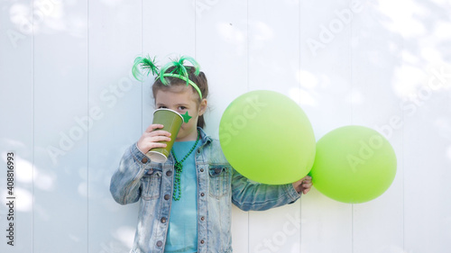 Photography Little kid with green funny horns and carnival star, holds two green balloons, drinks tea from paper cup and looks at the camera, celebrating saint patrick's day