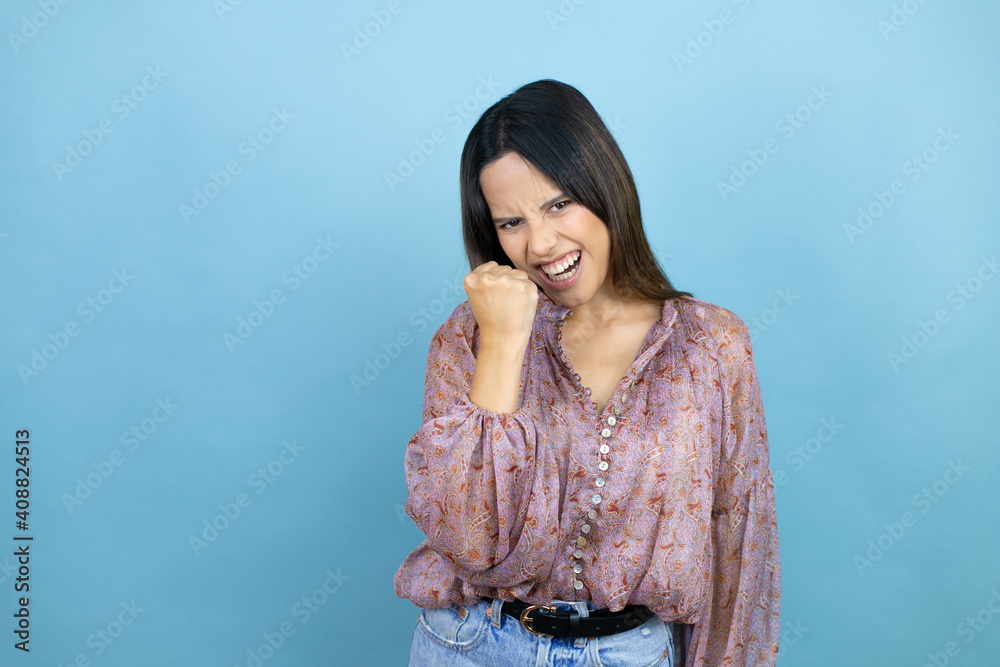 Beautiful latin woman wearing a pink shirt over blue isolated background angry and mad raising fist frustrated and furious while shouting with anger. Rage and aggressive concept. Beautiful latin woman wearing a pink shirt over blue isolated background angry and mad raising fist frustrated and furious while shouting with anger. Rage and aggressive concept.