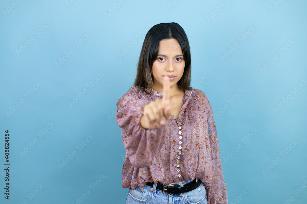 Beautiful latin woman wearing a pink shirt over blue isolated background showing and pointing up with fingers number one while is serious