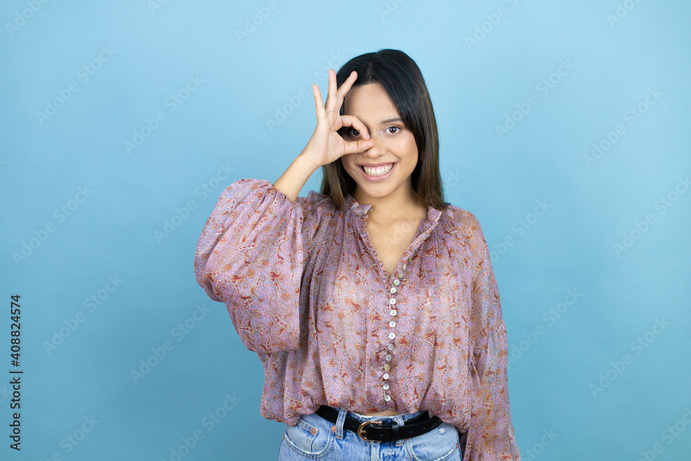 Beautiful latin woman wearing a pink shirt over blue isolated background doing ok gesture shocked with smiling face, eye looking through fingers Beautiful latin woman wearing a pink shirt over blue isolated background doing ok gesture shocked with smiling face, eye looking through fingers