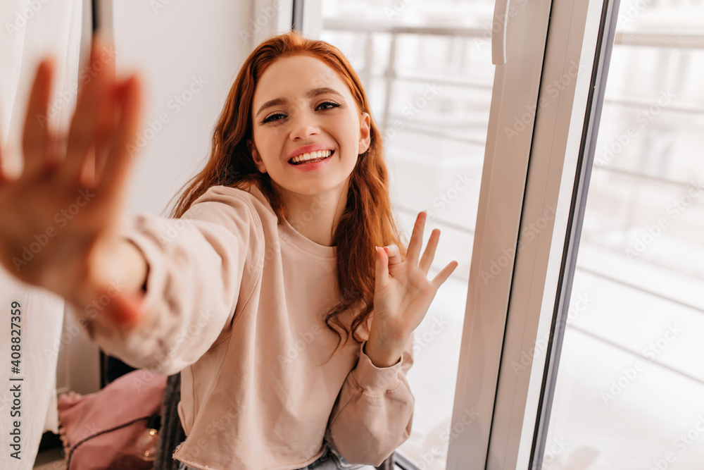 Good-humpured ginger girl posing near window. Indoor photo of laughing elegant lady making selfie. Good-humpured ginger girl posing near window. Indoor photo of laughing elegant lady making selfie.