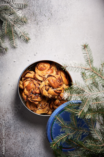 Freshly made Swedish swirl buns with cinnamon, sprinkled with pearled sugar, in tin cookie box with lid open. Light background with fir tree branches slightly overlapping the box