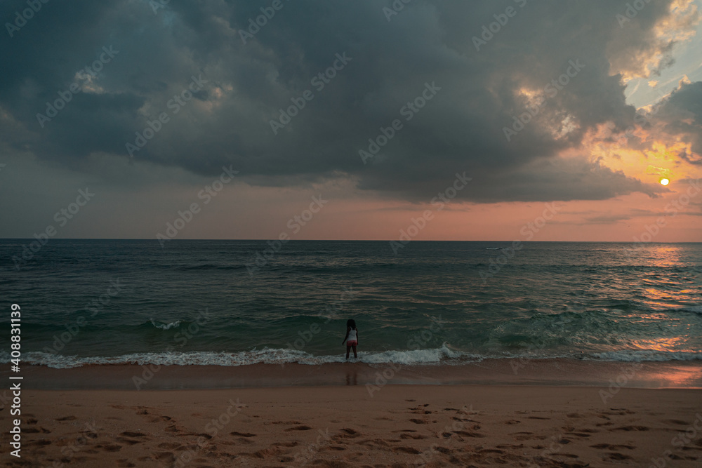 Kobieta na plaży na tle oceanu, zachodzącego słońca i pochmurnego nieba.