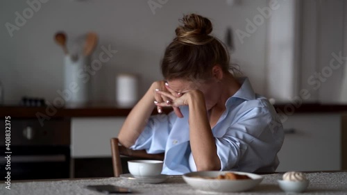 a blonde woman in a white top and blue shirt is sitting at the kitchen table. she puts face down on her clasped hands, then raises head. side view. the average plan