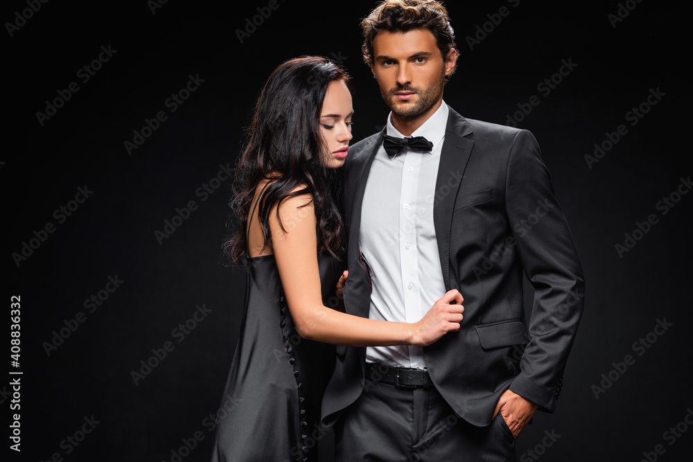 confident man with hand in pocket looking at camera near seductive woman in satin dress isolated on black