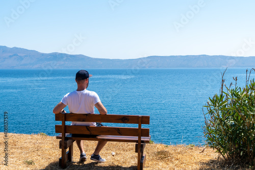 Guy sitting on wooden bench and looking at the blue ocean. Balos close to Chania. Grece