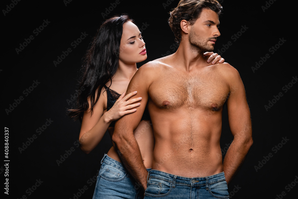 seductive brunette woman hugging muscular man looking away isolated on black seductive brunette woman hugging muscular man looking away isolated on black
