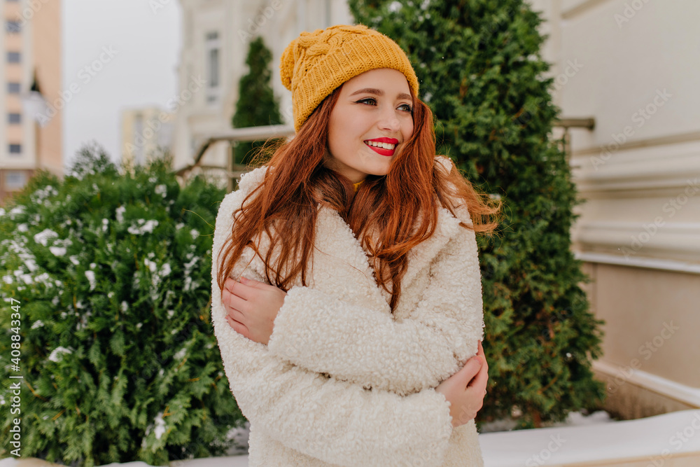 Blithesome female model in white coat posing in cold day. Outdoor shot of winsome ginger lady standing on the street. Blithesome female model in white coat posing in cold day. Outdoor shot of winsome ginger lady standing on the street.