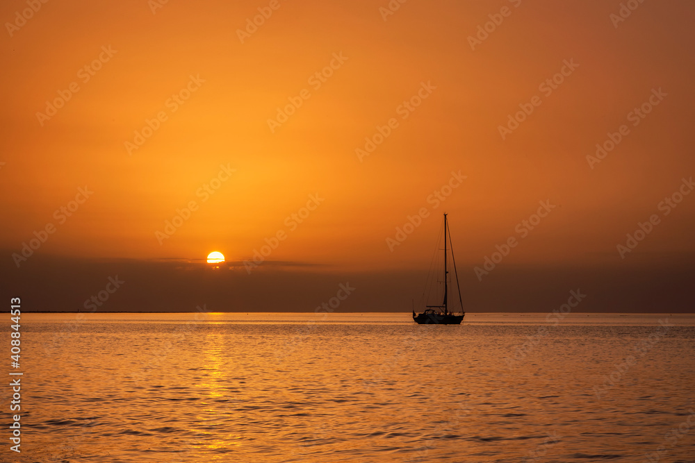 Sailing boat anchored at sea drenched in a golden sunset with sun just above horizon