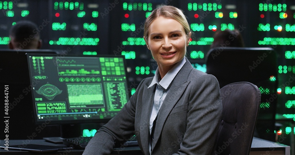 Blonde female programmer sitting at computer in server room working with database returns to camera and smiling. System software concept. Blonde female programmer sitting at computer in server room working with database returns to camera and smiling. System software concept.