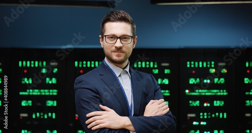 Successful male programmer in jacket and glasses standing on background of running servers looking at camera with his arms crossed.