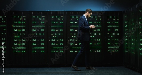 Smart computer technician walks along the cabinets with servers holding tablet, and monitors operation of devices in server room.