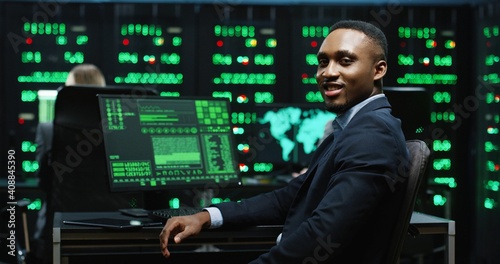 African-American IT worker sitting in server room working with database on computer returns to camera and smiling. System software concept.