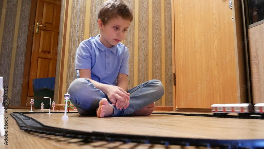A cute boy 6-7 years old plays with a children's railway, sits on the floor and looks at the traveling trailers A cute boy 6-7 years old plays with a children's railway, sits on the floor and looks at the traveling trailers