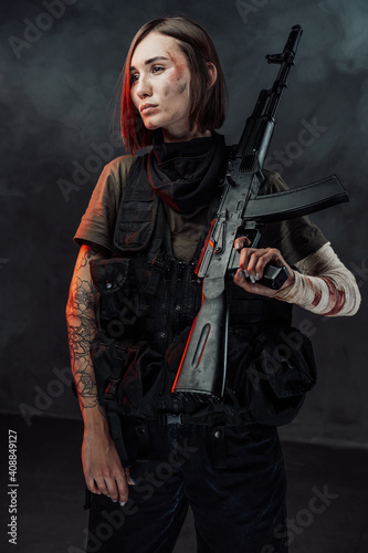 Seductive female soldier with tattoo and bandaged hand poses in dark foggy background holding rifle on her shoulder.
