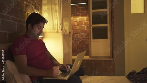 shot of a young Indian man sitting at home busy working on a laptop while his wife surprises him with a cake and both celebrates birthday by blowing up candle light and eats it