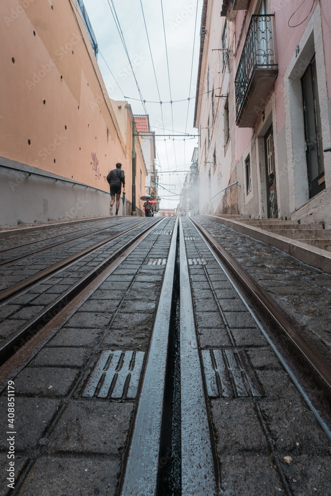 Fototapeta premium Photograph of a runner from the back, jogging on a steep street in Lisbon, where the tram rails that run through the city can be seen on the ground.