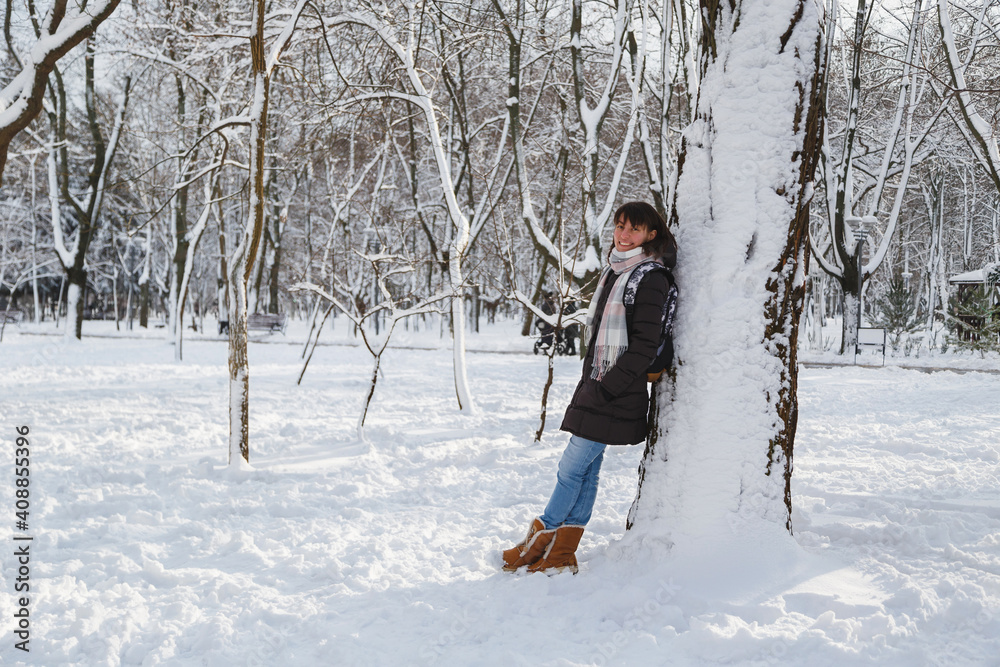 Young smiling woman standing near to a big tree in a snow-covered winter park. Snowy winter
