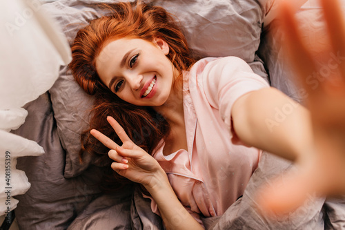 Relaxed red-haired girl lying in bed with charming smile. Indoor photo of sensual female model making selfie in bedroom.